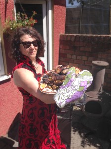 Plate of grub, green and mauve original Art Nouveau suffragette designed oven gloves held by smiling woman in dark glasses in red dress 