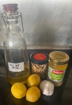 A clear bottle of extra virgin olive oil, dried chickpeas in a jar, jar of tahini (brand: Bodrum organic), 3 lemons and 1 bulb of garlic.